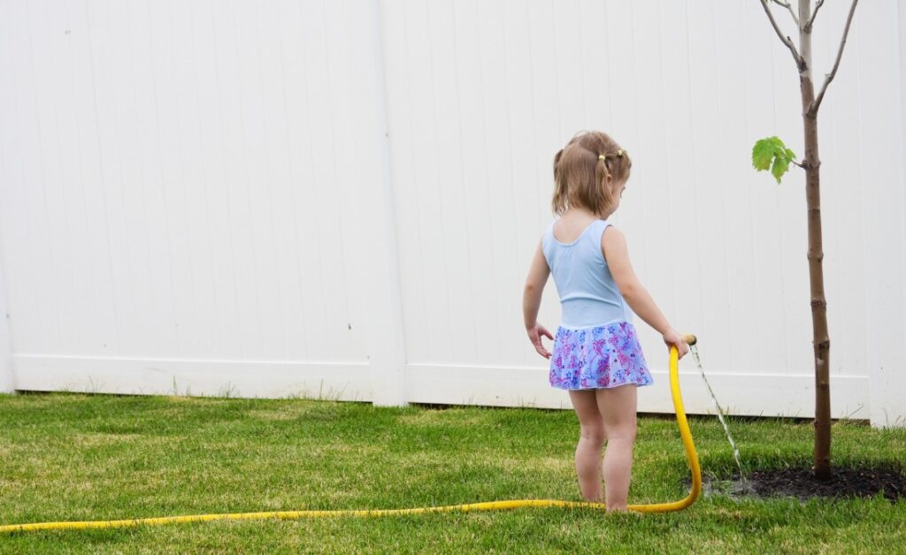 A young girl watering a newly planted tree with a garden hose set to a slow drip in a Cedar Rapids backyard.