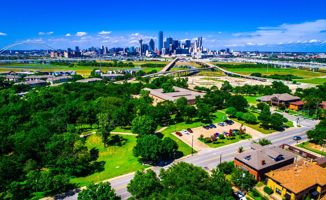 An aerial view shows a Dallas neighborhood filled with mature trees in the foreground, with the downtown skyline visible in the distance under a bright blue sky.