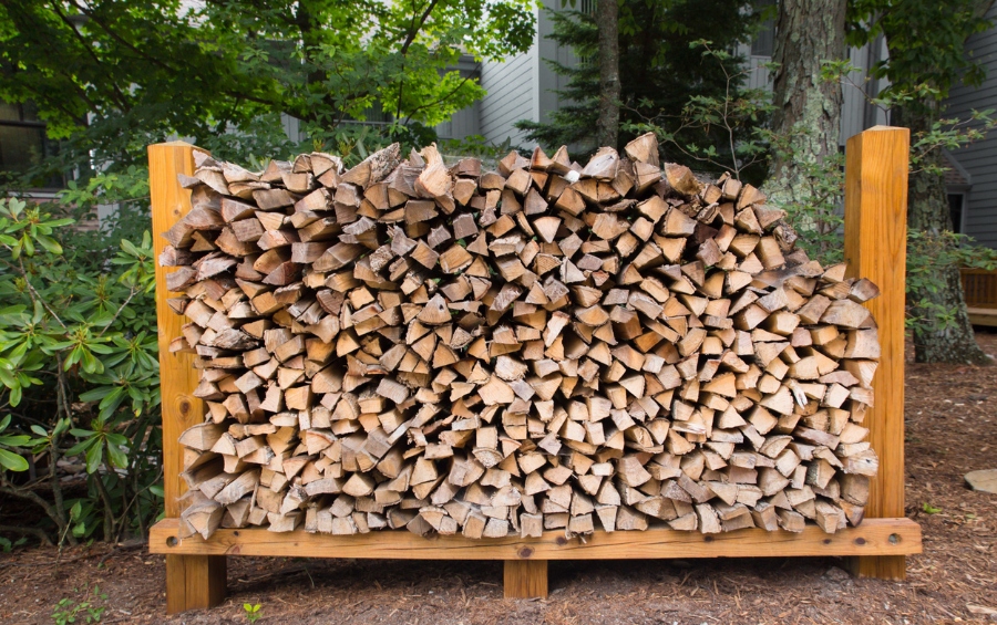 A large rack of neatly stacked split firewood sitting on mulch in a residential backyard with green trees and a house visible in the background.