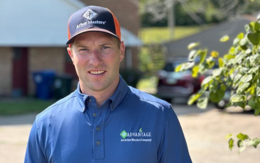 Closeup of a smiling Arbor Masters arborist wearing company branded navy blue polo shirt and orange and navy baseball cap standing outdoors near residential property with trees in background