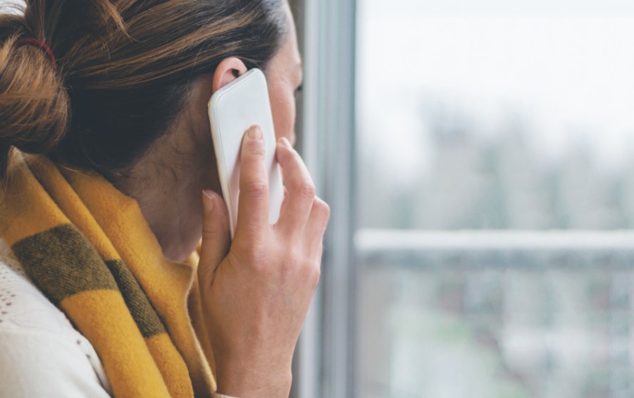 A woman holding a phone to her ear while looking out a window on an overcast day, wearing a cozy yellow and brown striped scarf.