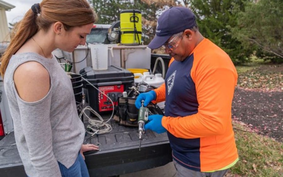 An Arbor Masters plant health care technician prepares a tree injection unit at the truck tailgate while a homeowner looks on.