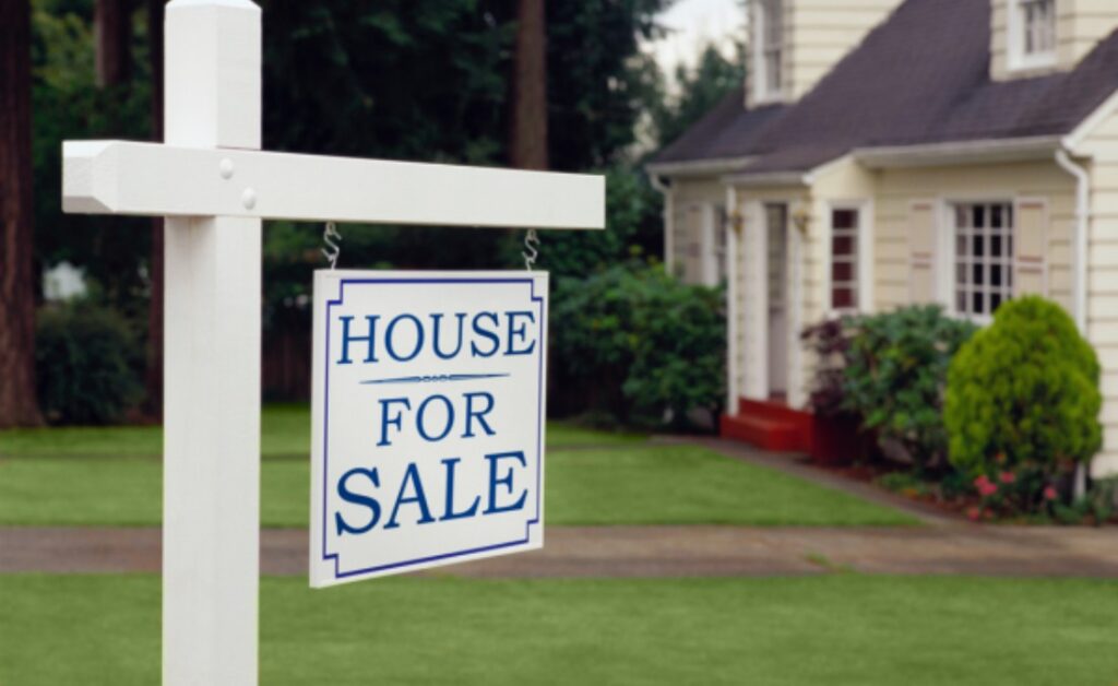 A white "House For Sale" sign with a blue border stands on a lawn in front of a light yellow house.