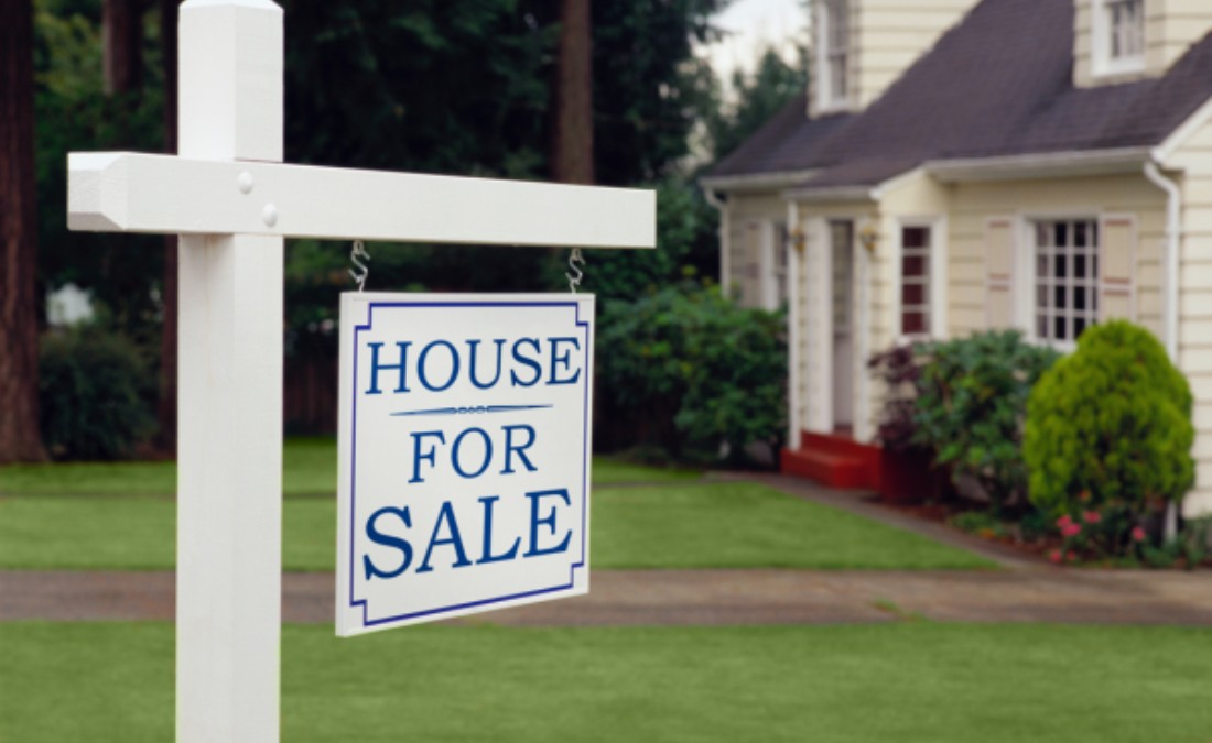 A white "House For Sale" sign with a blue border stands on a lawn in front of a light yellow house.