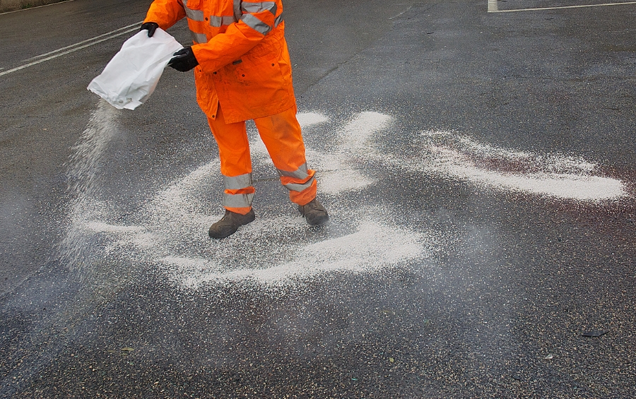 A public works employee in orange safety gear spreads road salt on an asphalt street during winter operations.