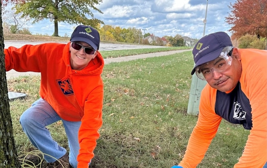 Two Arbor Masters ISA Certified Arborists kneel at the base of a tree to perform a plant health care assessment, examining soil conditions and root structure.