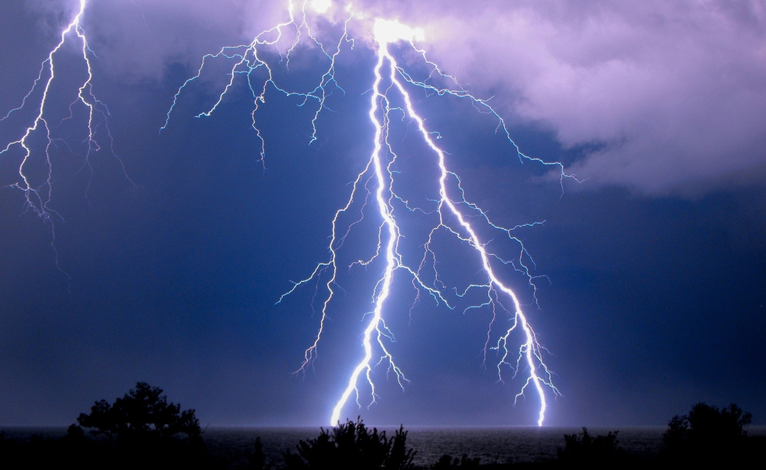 Lightning bolts striking during a severe thunderstorm over a tree line at night.