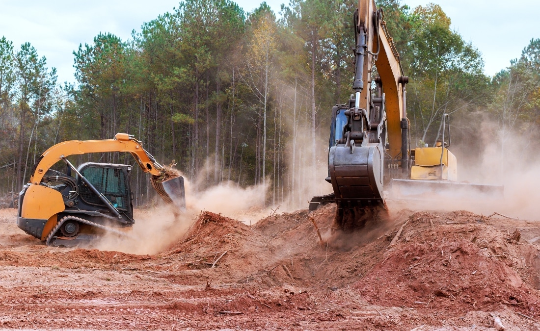 A skid-steer loader and a tracked excavator working together to clear a wooded Missouri lot, kicking up a haze of red clay dust against a backdrop of standing pine trees, with cleared soil and root debris in the foreground.