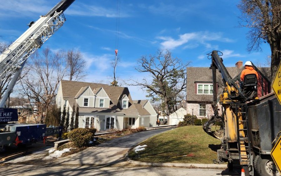 Crane lifting a large tree section over a residential home while an operator in orange safety gear guides the load from a grapple truck.