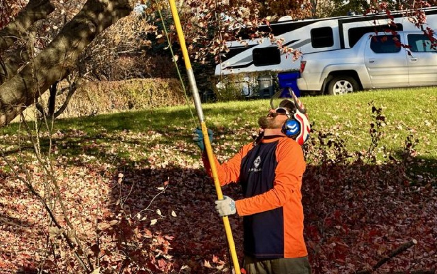 Arbor Masters arborist in an orange safety shirt and hearing protection using a pole saw to prune branches in a residential Kansas City yard. 