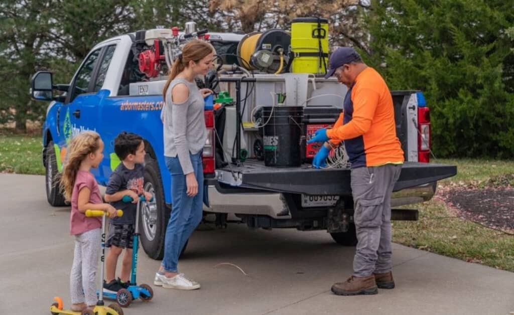 Arbor Masters plant health care technician showing equipment from the back of a service truck to a mom and two young kids in a residential driveway.