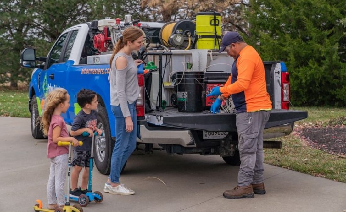 Arbor Masters plant health care technician showing equipment from the back of a service truck to a mom and two young kids in a residential driveway.