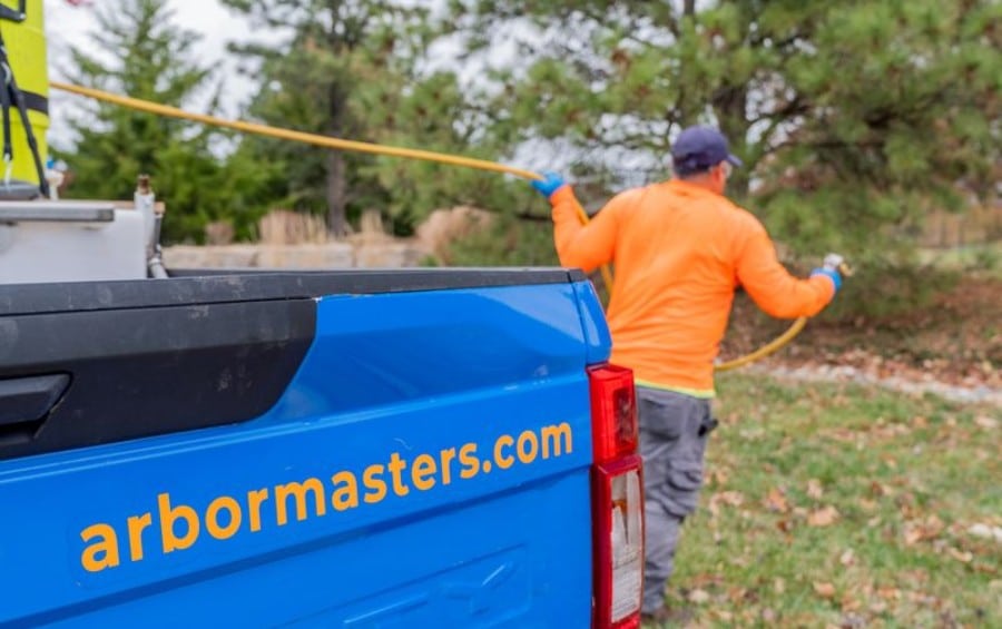 Arbor Masters technician in an orange long-sleeve shirt and blue gloves spraying from a hose connected to a branded service truck in a residential yard.