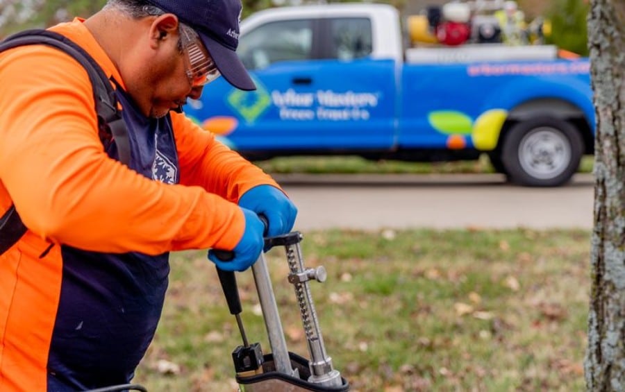 Arbor Masters technician in safety glasses and blue gloves operating a tree trunk injector at the base of a tree, with a branded service truck in the background.
