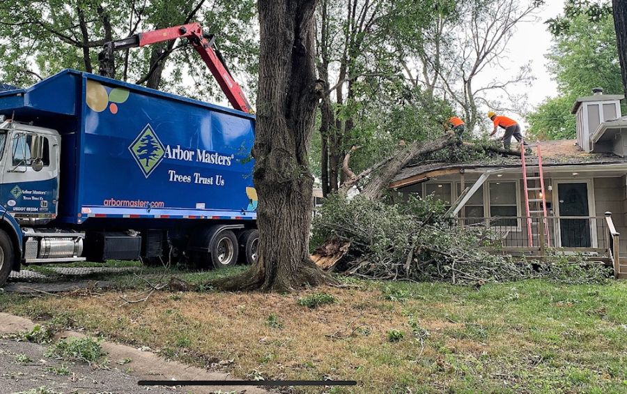 Arbor Masters on site in Raytown, MO, cleaning up storm debris after a large tree fell onto a home, causing significant damage. The team is loading fallen branches and limbs into a large truck for removal and disposal.