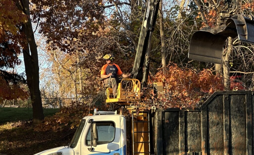 An Arbor Masters blue and white bucket truck with extended boom operates on a street while crew members manage tree debris cleanup operations.