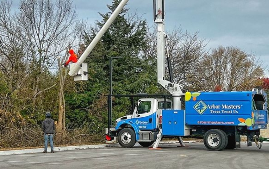 An Arbor Masters crew member in safety gear operates from a yellow bucket truck lift to reach high branches while working near a residential fence and grapple truck.