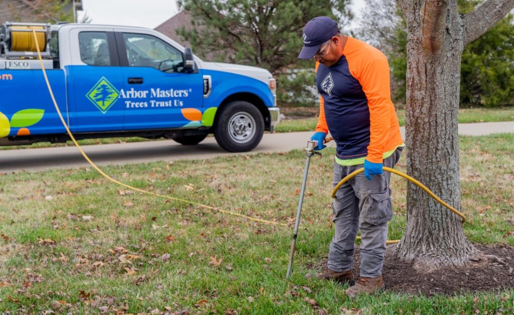 An Arbor Masters tree care professional in orange safety clothing injecting fertilizer around the root zone of a tree, with the company's blue truck visible in Wichita.
