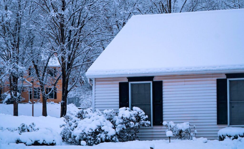 A residential home with a snow-covered roof and landscaping featuring mature trees in a winter neighborhood setting.