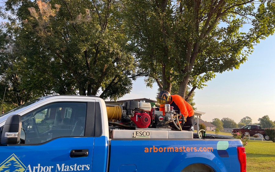 An Arbor Masters technician in an orange shirt and helmet operating a commercial sprayer mounted in the bed of a blue Arbor Masters pickup truck parked beneath a large shade tree.