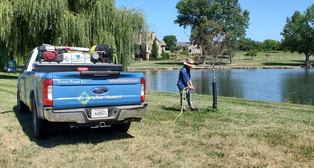 An Arbor Masters technician operates fertilization equipment near a young tree while an Arbor Masters truck with specialized equipment sits parked on the lawn beside a residential pond.